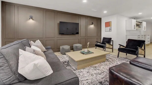 Ein modernes Wohnzimmer mit einem grauen Sofa, einem Couchtisch und zwei Sesseln. Ein Flachbildfernseher ist an der Wand befestigt, und im Hintergrund ist eine Küchenzeile zu sehen.