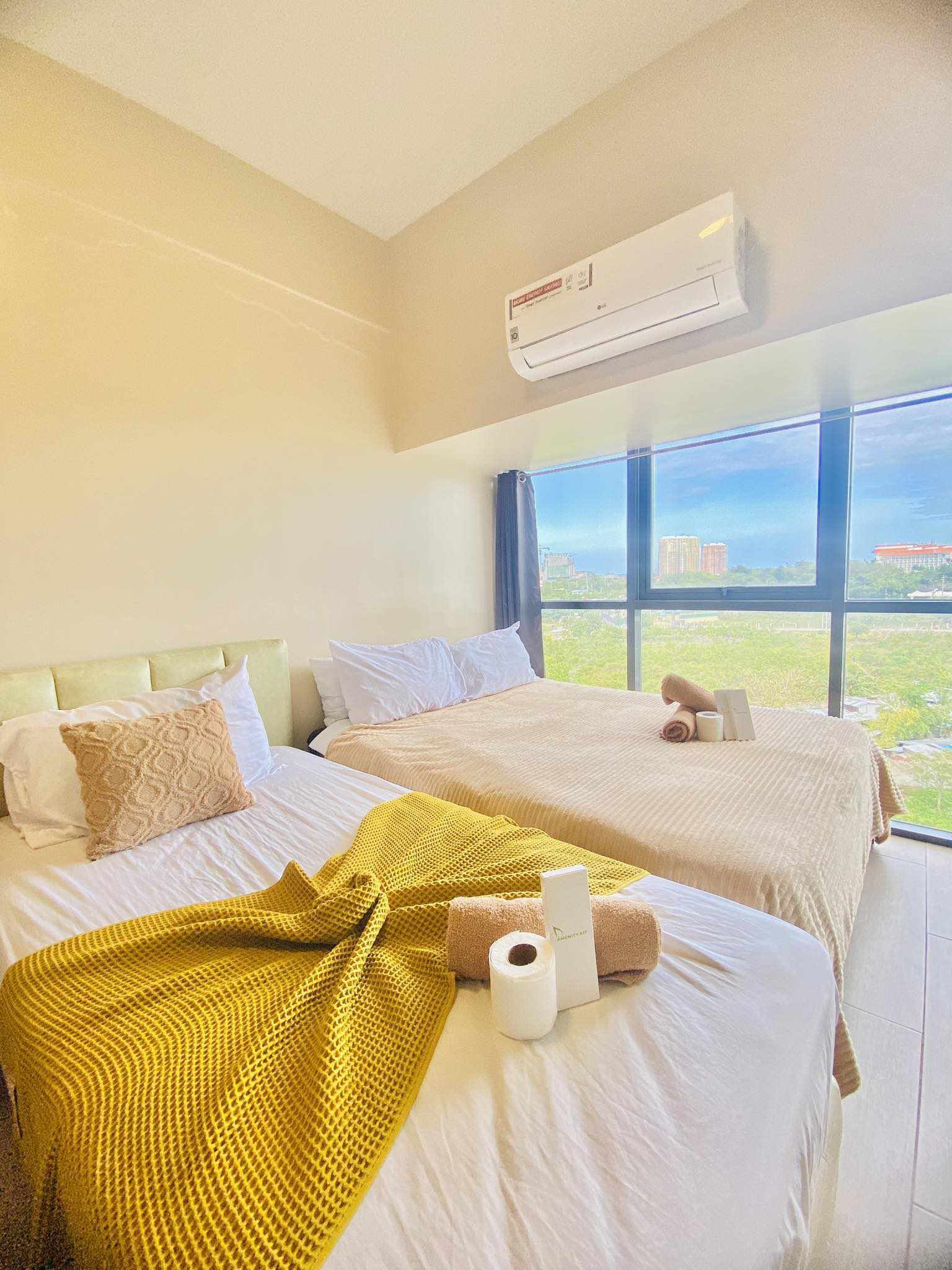 A hotel room with two beds, a large window with a city view, an air conditioning unit, and a yellow blanket on one bed.