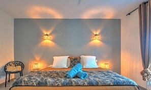 A bedroom with a bed, two wall-mounted lights, a chair, and a decorative item on the wall. - Beautiful 2-bedroom beachfront apartment in Grand Case (Saint-Martin)