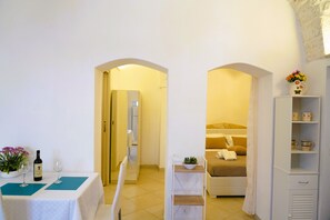 A hotel room with a bed, a bedside table with a vase of flowers, a wardrobe, and a table with a bottle of wine and glasses. - La Casetta in Pietra - Arcuève (Ostuni)