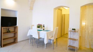 A dining area with a table set for two, a television on a stand, and a wooden shelf with decorative items. - La Casetta in Pietra - Arcuève (Ostuni)