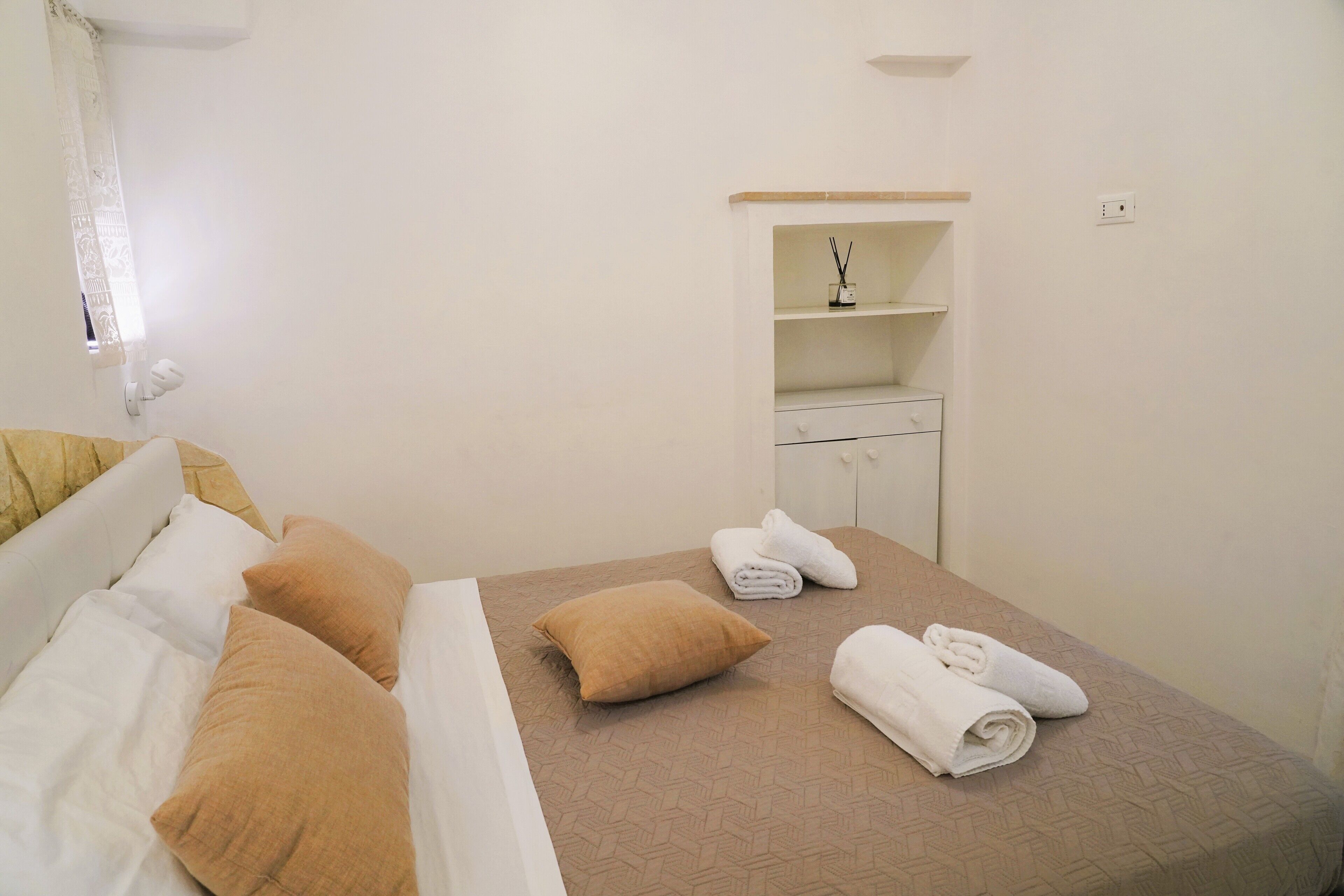 A hotel room with a bed, pillows, a small shelf with toiletries, and a cabinet.