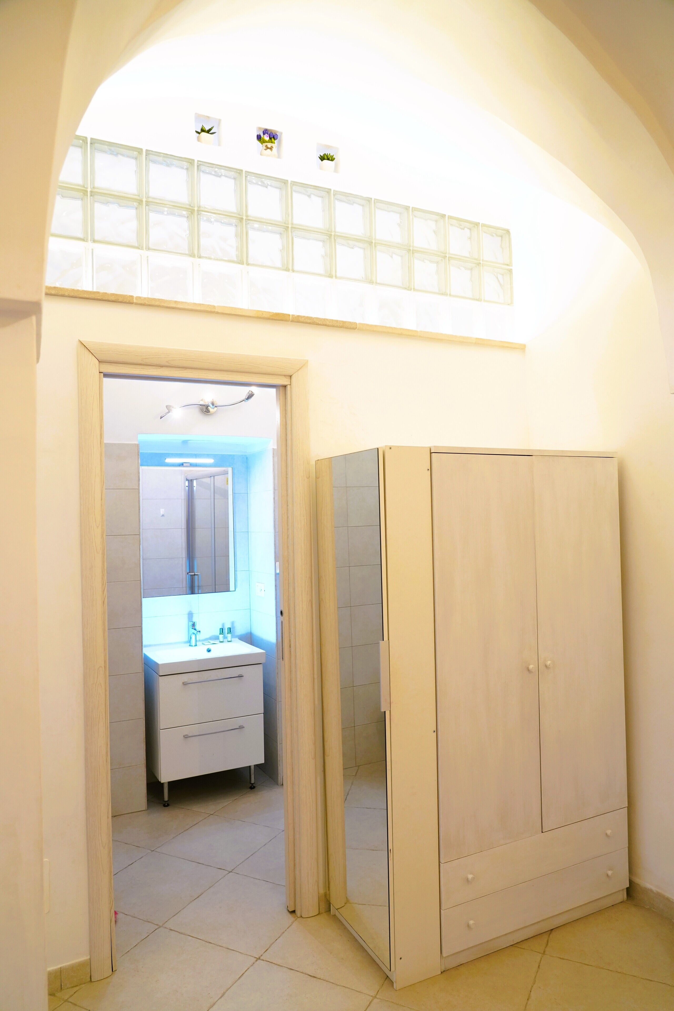 Un pasillo con un armario de madera, un mueble blanco con espejo y piso de baldosas.