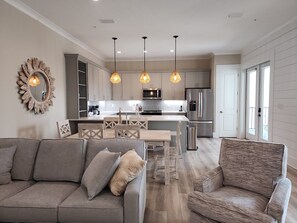 A modern kitchen with a central island, pendant lighting, and a dining area with a wooden table and chairs. - Beach Daze - Short walk to the beach and restaurants! (Gulf Shores)