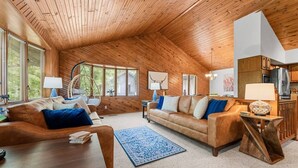 A cozy living room with wooden walls and ceiling, a brown leather sofa, a blue rug, and a dining table.