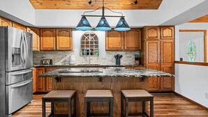 A kitchen with wooden cabinets, a stainless steel refrigerator, a large island with stools, and a unique window design.