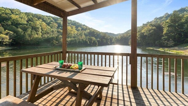 A wooden deck with a table set for two, overlooking a river and forested hills.