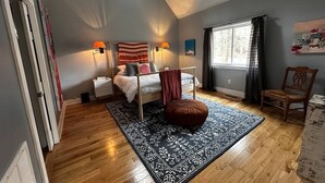 Un dormitorio con techo inclinado, un ventanal con cortinas, una cama con almohadas, una mesita de noche, una silla y una alfombra.