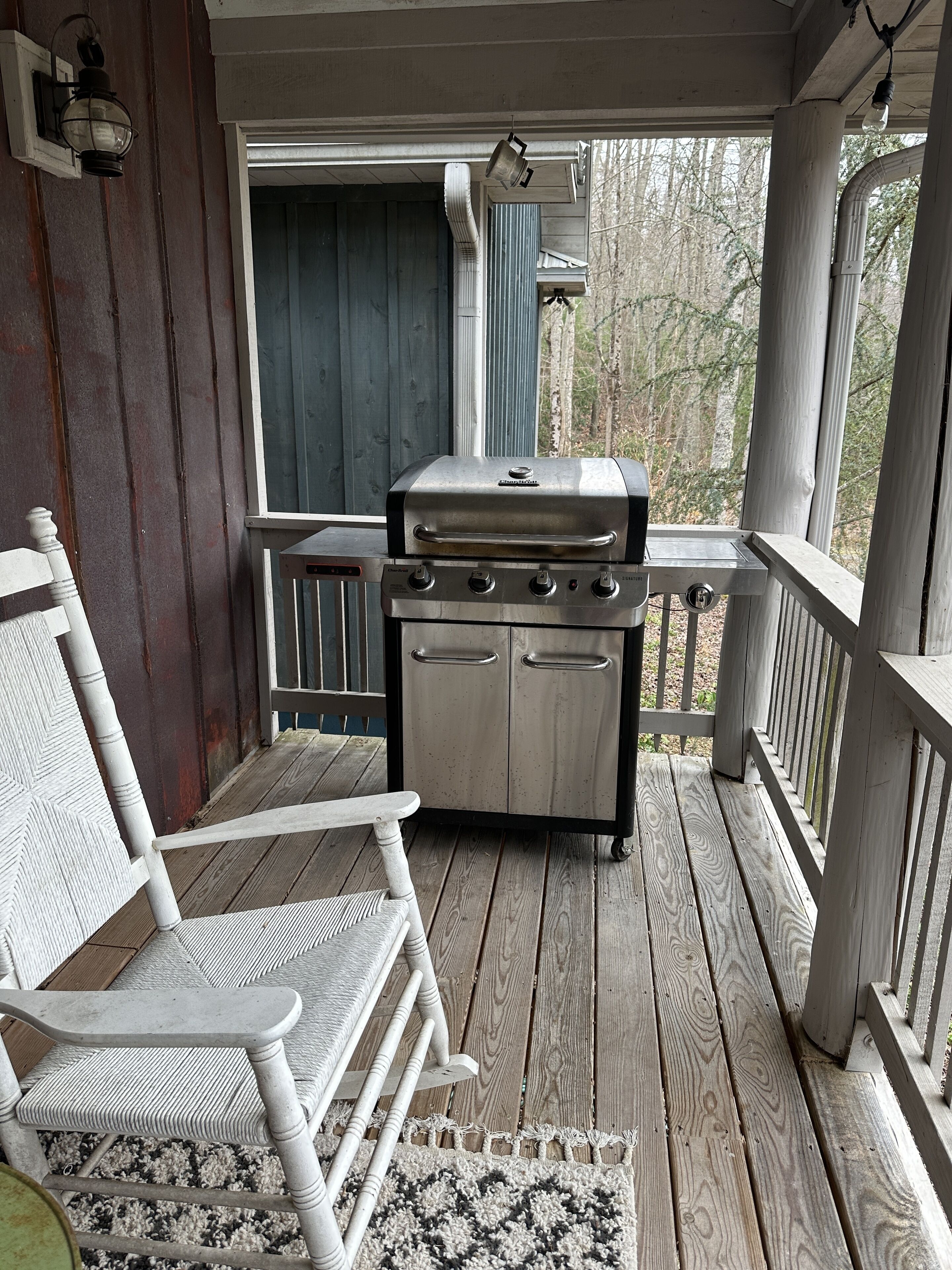 A wooden porch with a grill, a white chair, and a view of trees.