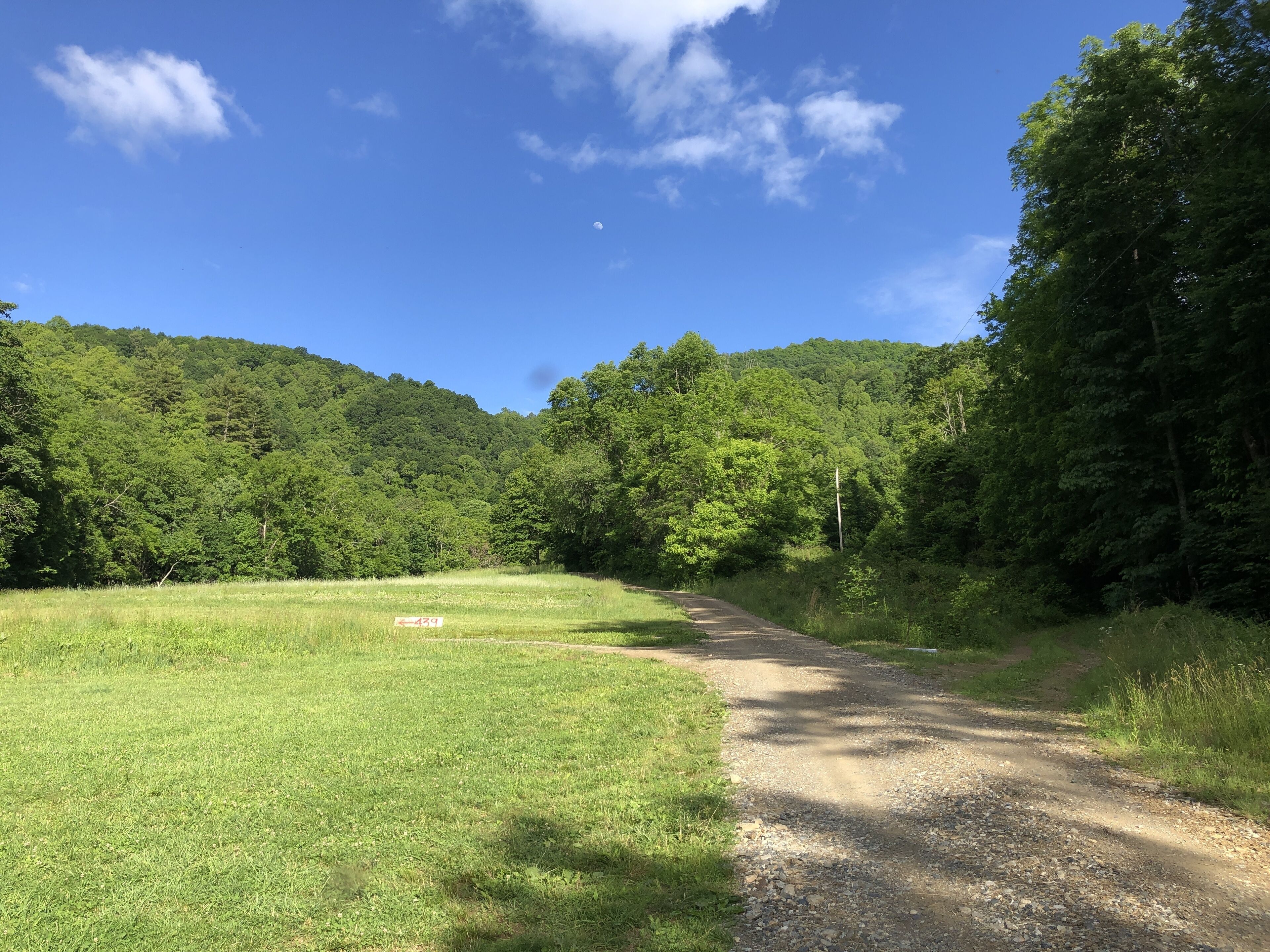 A rural road leading through a green field with a forested hill in the background.