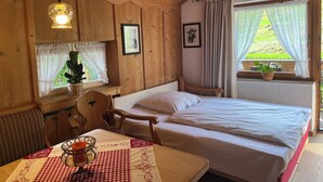 Une pièce chaleureuse aux murs et au plafond en bois, un lit avec une literie blanche, une table recouverte d’une nappe à carreaux, une chaise et une fenêtre avec des rideaux.