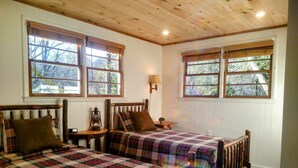 Una habitación acogedora con dos camas de madera, una mesita de noche con lámpara, y ventanas amplias con vistas a árboles.
