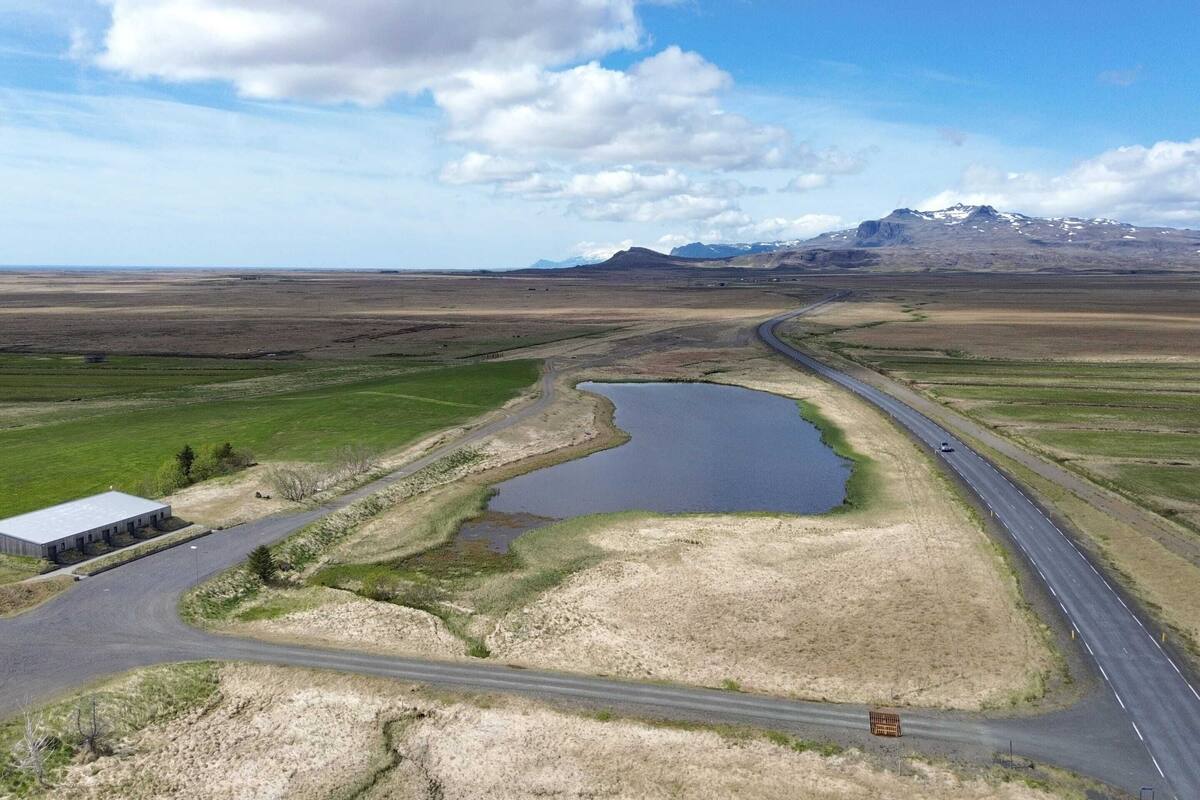 A rural landscape with a road curving around a small body of water, a building on the left, and mountains in the background.