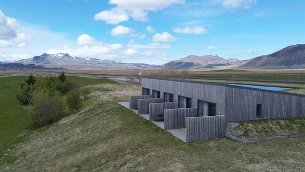 A modern building with a flat roof and large windows, situated on a grassy hill with a mountain range in the background. - Eiðhús Apartments (Eyja- og Miklaholtshreppur)