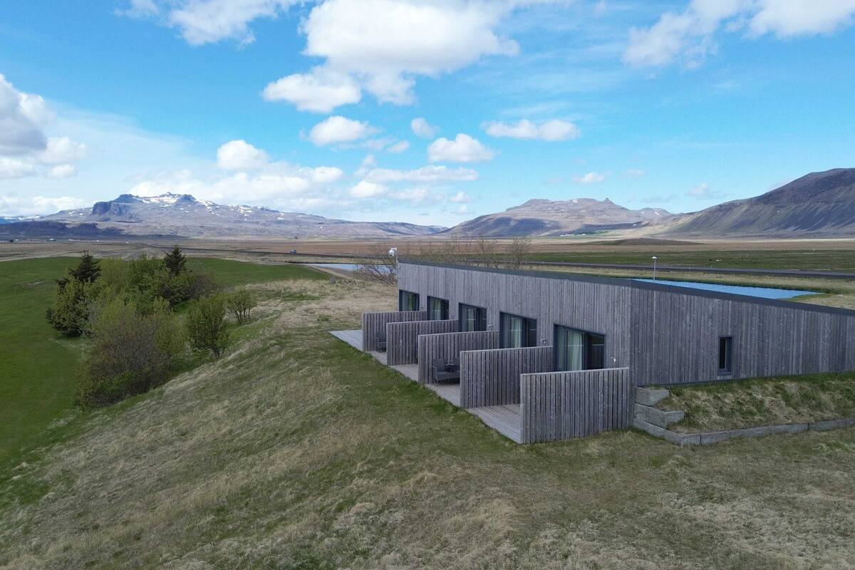 A modern building with a flat roof and large windows, situated on a grassy hill with a mountain range in the background.