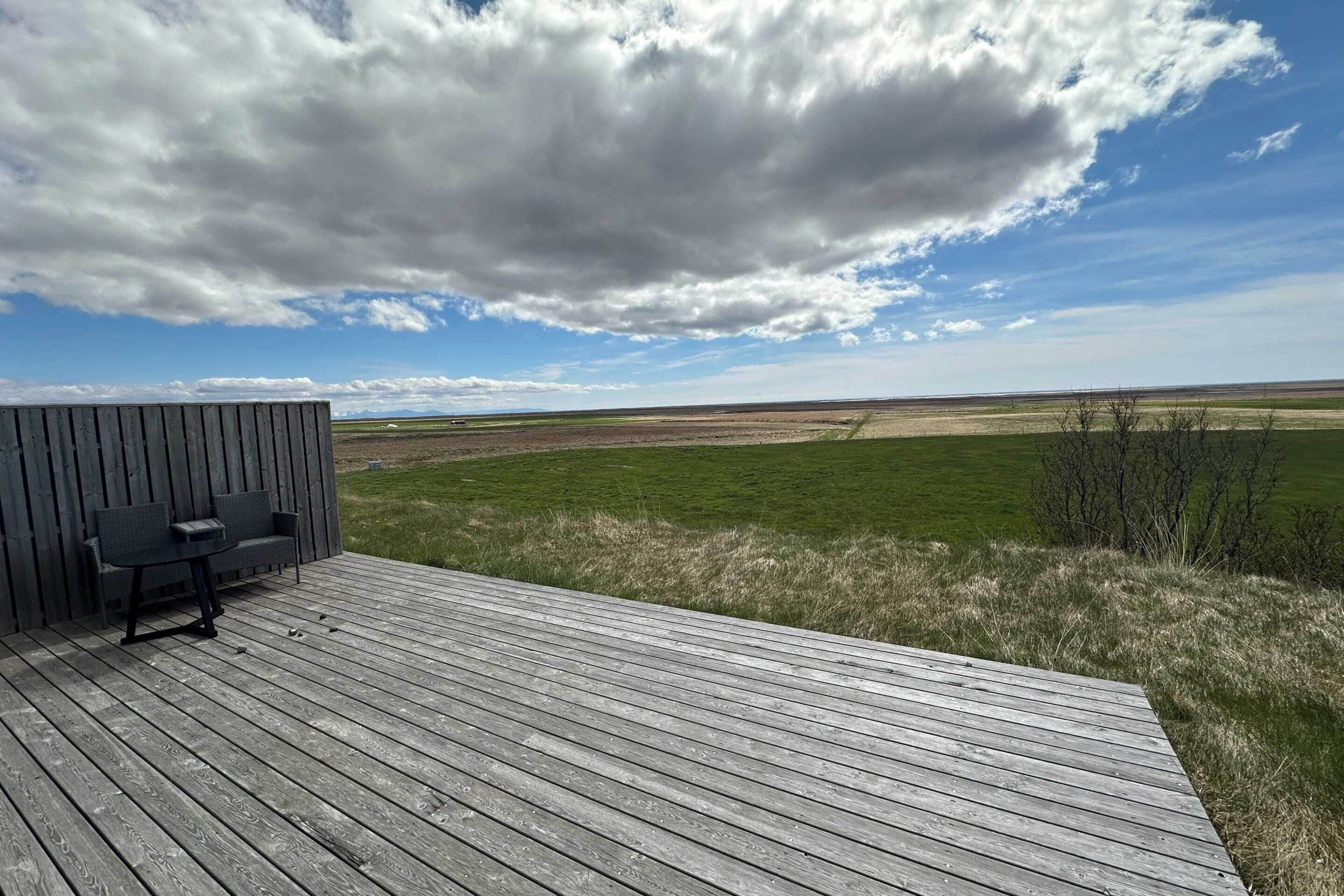 A wooden deck with a bench and table, overlooking a grassy field and distant hills under a partly cloudy sky.