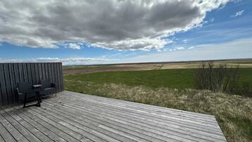 A wooden deck with a bench and table, overlooking a grassy field and distant hills under a partly cloudy sky.