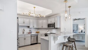 A modern kitchen with white cabinetry, a central island with stools, stainless steel appliances, and a unique chandelier. - Serenity Awaits: Unwind in Style with Our Premier Orange Beach Condo Rental (Orange Beach)