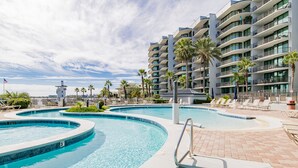 A hotel with a large, curved swimming pool surrounded by lounge chairs and palm trees. - Serenity Awaits: Unwind in Style with Our Premier Orange Beach Condo Rental (Orange Beach)