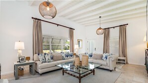 Un salon spacieux avec une table basse en verre, deux canapés ornés de coussins à motifs, une table d’appoint avec une lampe et une grande fenêtre avec des rideaux.