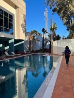 Una piscina con sillas de descanso, palmeras y un edificio moderno con ventanas amplias.
