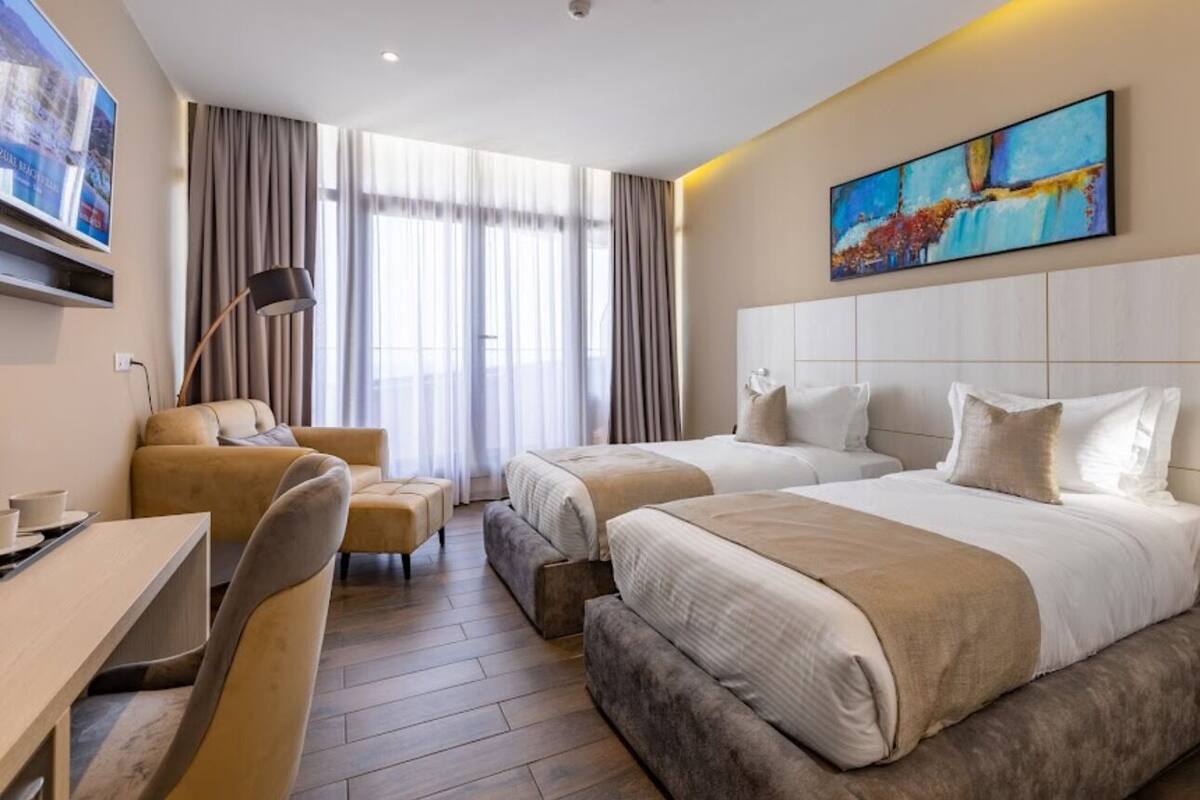 A hotel room with two beds, a desk, a chair, and a large window with curtains.
