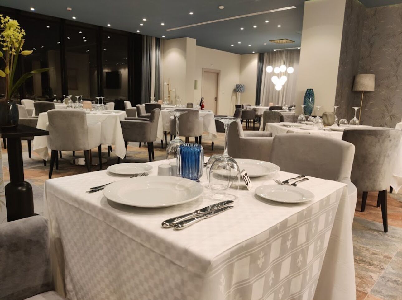 A hotel dining area with multiple tables set for dining, each with white tablecloths and silverware.