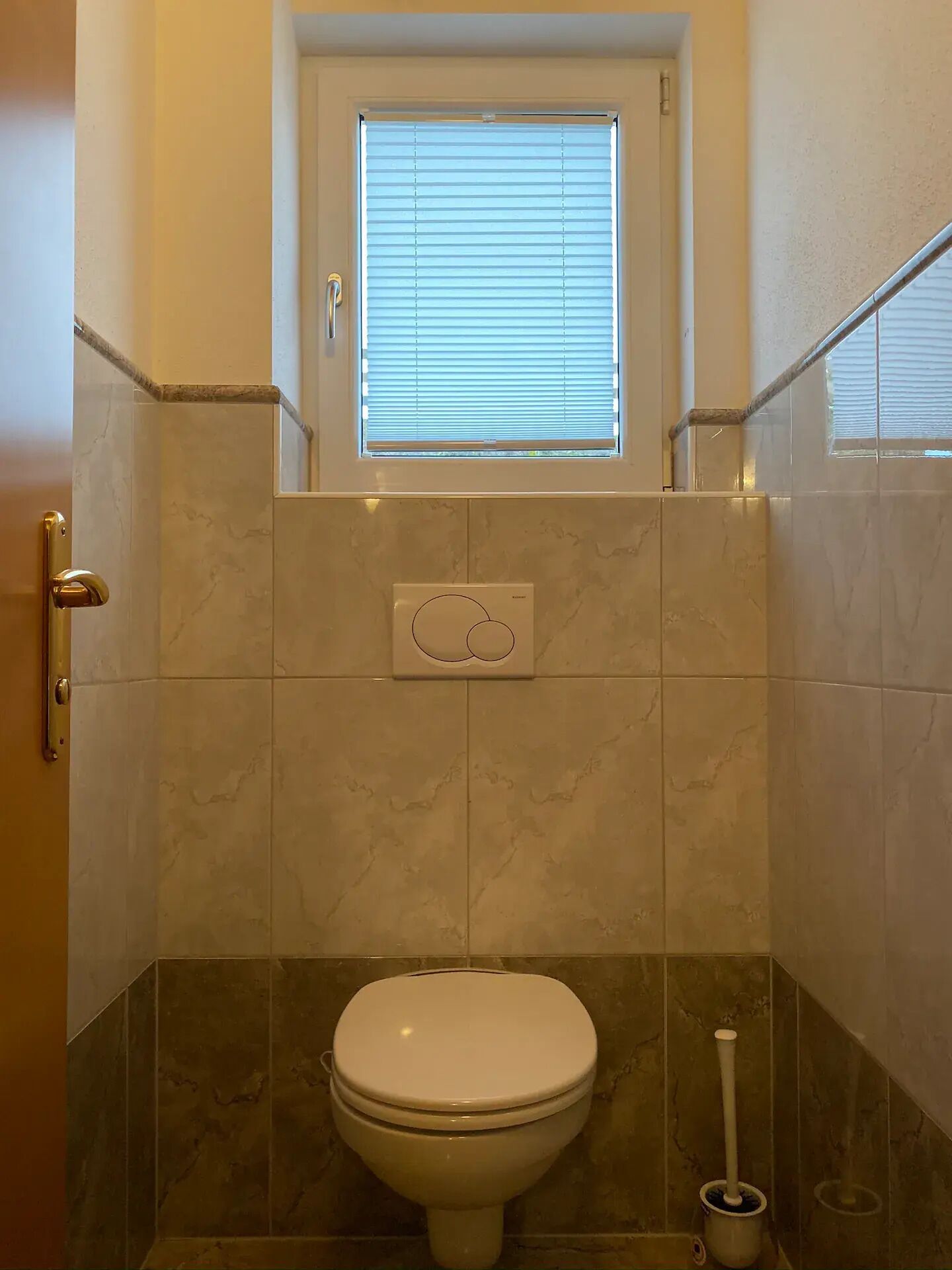 A bathroom with a toilet, a window above it, and a wall-mounted flush handle.