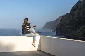A person sitting on a white balcony overlooking the ocean with a cliff in the background. - On the North Coast, by the sea - Casa do Terreiro (São Vicente)