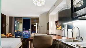 A hotel room with a bed, a desk, a chair, a kitchen area, and a view of the cityscape.