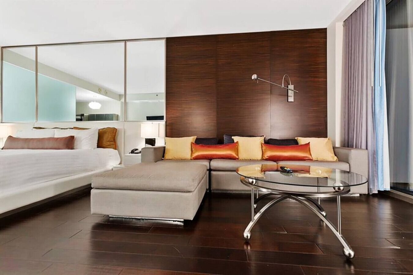A modern hotel room with a large bed, a sofa, a glass coffee table, and a large mirror.