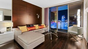 A modern hotel room with a sofa, a glass table, a view of the cityscape, and a wooden accent wall.