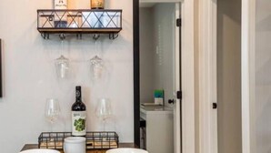 Un comedor con una mesa dispuesta para cuatro personas, dos botellas de vino en una estantería superior y un refrigerador al fondo.