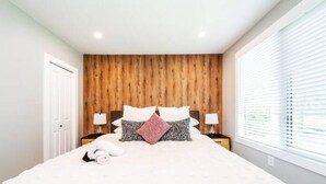 Un dormitorio con cabecera de madera, una cama con ropa de cama blanca, dos mesitas de noche con lámparas y una ventana con persianas.