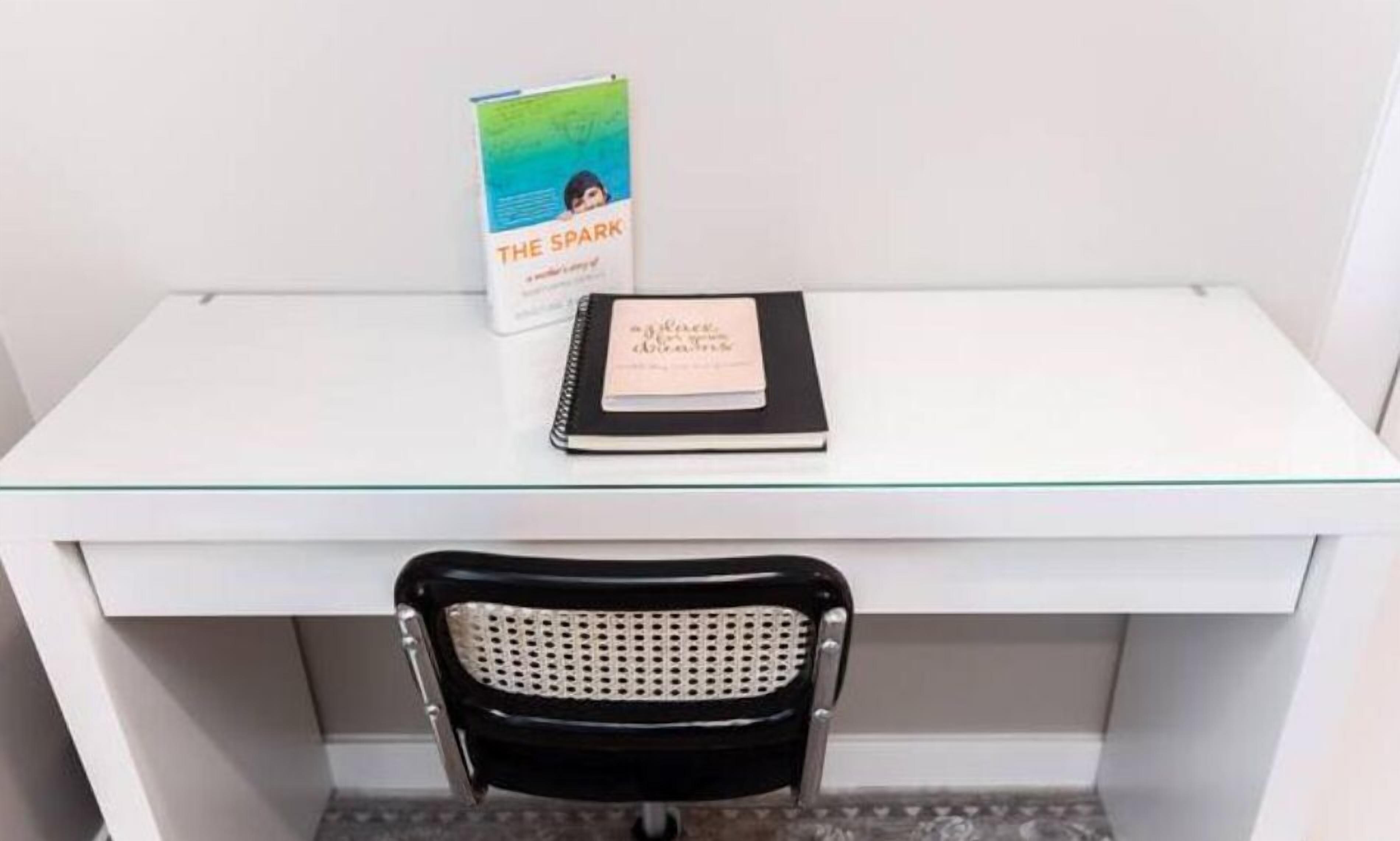 Een wit bureau met een glazen blad, een zwarte stoel met een geperforeerde rugleuning en een boek met de titel 'The Spark' op het bureau.