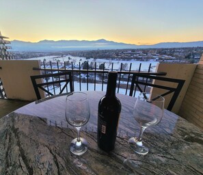 A table with a bottle of wine and two glasses on a balcony overlooking a snowy landscape with mountains in the distance.