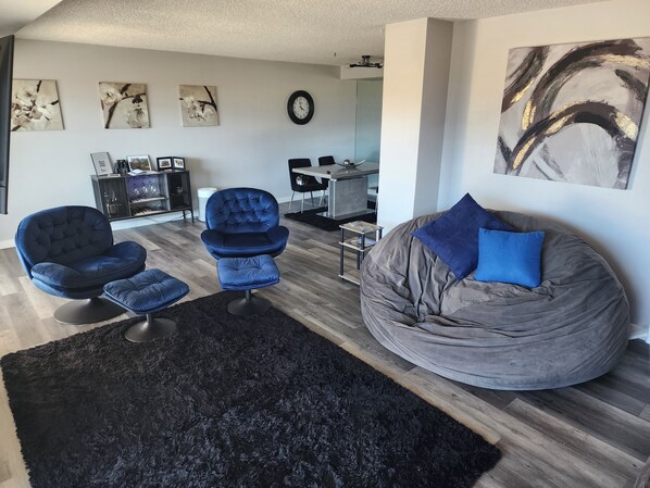 A modern living room with a black shag rug, a blue armchair, a grey bean bag, and a dining area in the background.
