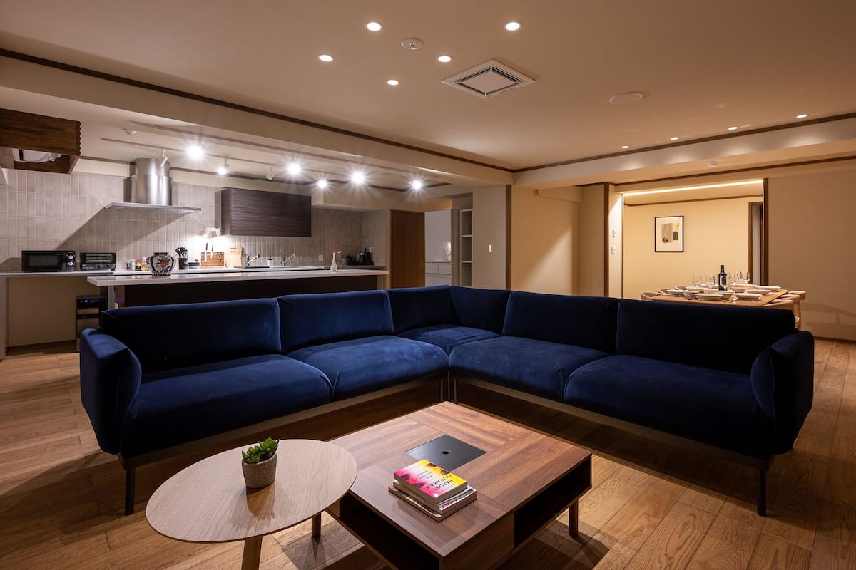 A modern living room with a dark blue sectional sofa, a round wooden coffee table with a book and a plant, and a kitchen area in the background.