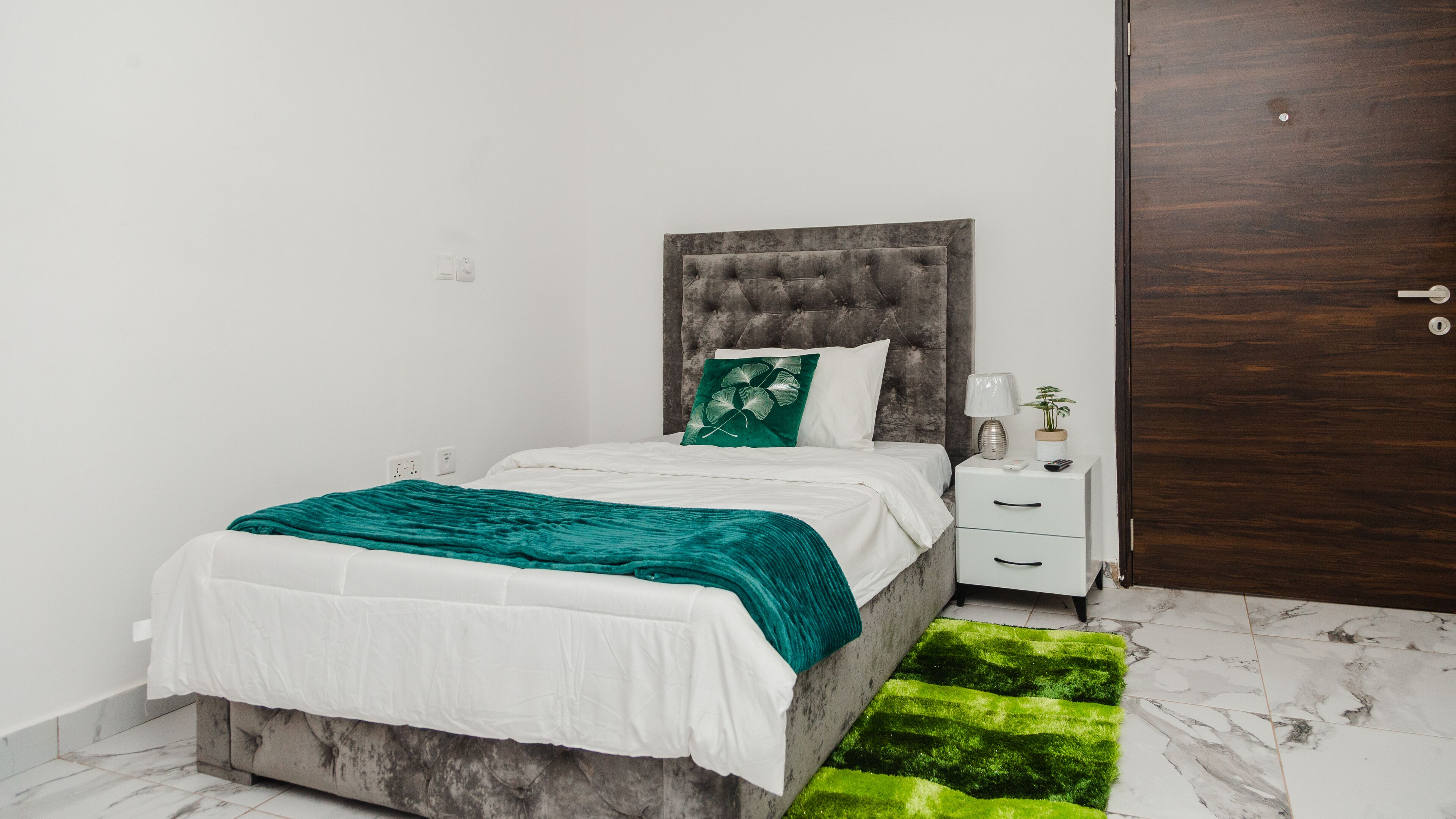 A bedroom with a bed, a nightstand, a lamp, a plant, and a wooden door.