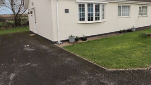 A single-story house with a tiled roof, white exterior walls, and a front porch. There are four windows on the side of the house, and several potted plants are placed along the front lawn. - 13 Gower Holiday Village (Swansea)