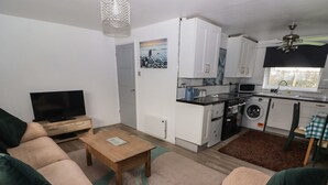A compact living space with a sofa, a coffee table, a TV, a kitchen with a washing machine, and a dining area with a table and chairs. - 13 Gower Holiday Village (Swansea)