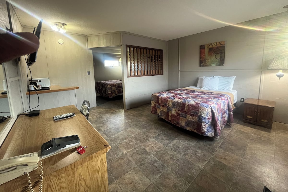 A hotel room with a bed, a desk with a telephone and remote control, a wall-mounted lamp, and a painting.