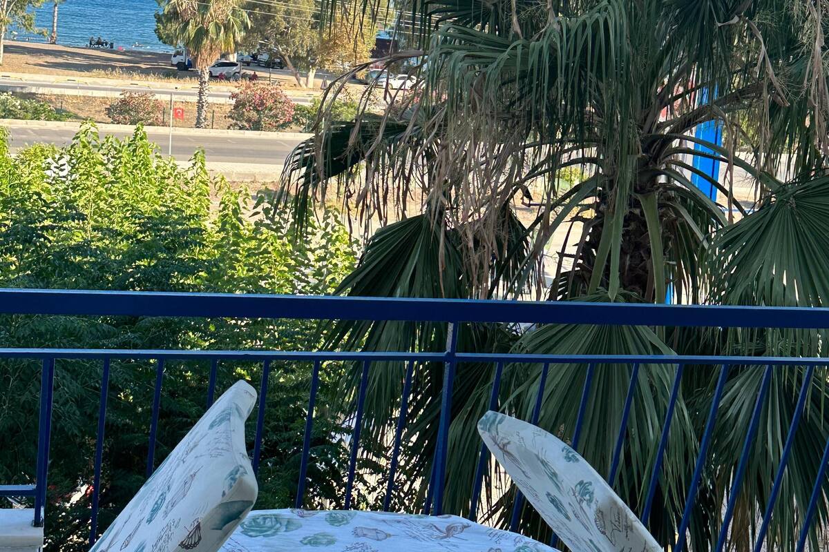 A balcony with a table covered in a floral tablecloth, overlooking a coastal area with palm trees and a view of the sea.