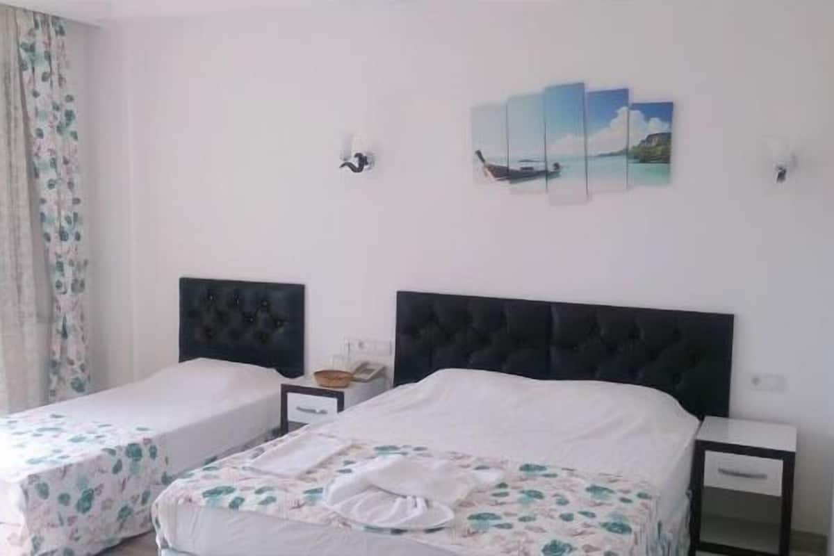 A hotel room with two single beds, a nightstand, a wall-mounted lamp, and a painting of a coastal scene.