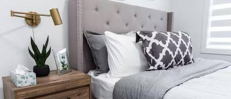 A neatly arranged bedroom with a tufted headboard, a bedside table with a lamp, a potted plant, and a framed picture.
