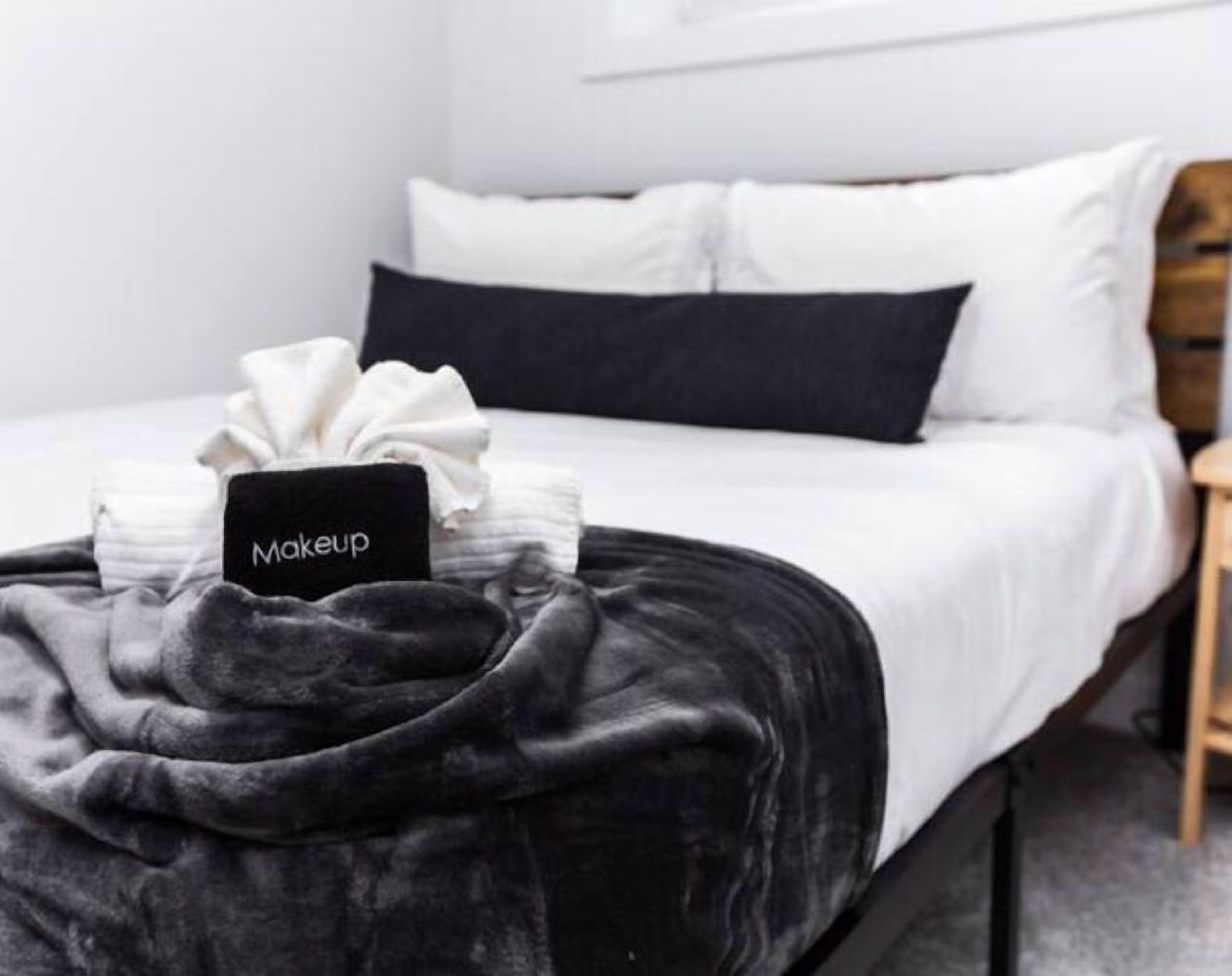 Ein ordentlich gemachtes Bett mit einer schwarzen Decke, weißen Kissen und einem Schild, das auf der Decke den Schriftzug „Makeup“ trägt.