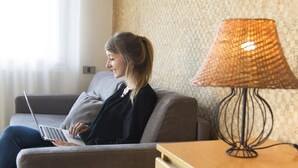Une personne est assise sur un canapé gris et utilise un ordinateur portable. Une lampe en osier est posée sur une petite table en bois à côté d’elle.