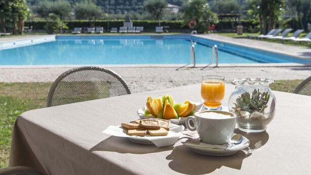 Une table avec des aliments pour le petit-déjeuner, une tasse de café et un verre de jus d’orange, le tout placé près d’une piscine avec vue sur les montagnes.