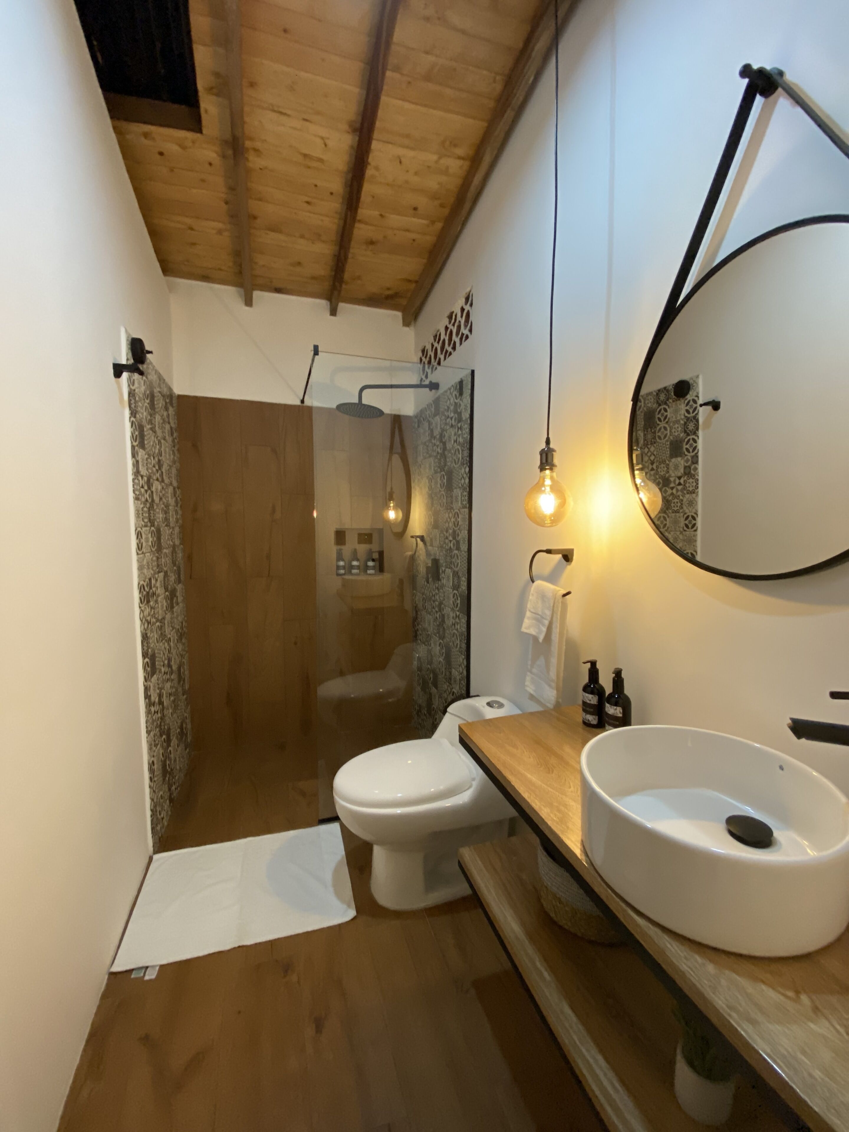 Un baño moderno con techo de madera, cabina de ducha con vidrio, inodoro blanco y tocador de madera con lavamanos redondo.
