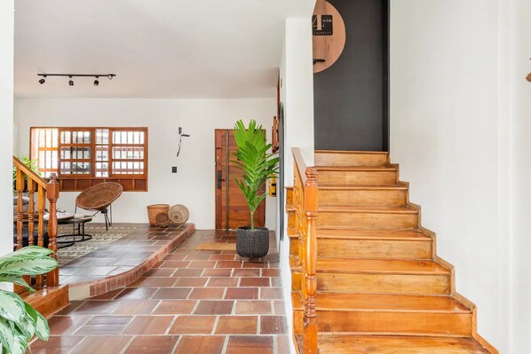 A staircase leading to a wooden door, a large potted plant, and a wicker chair in a room with tiled flooring and a window with wooden frames.